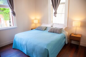 A neatly made bed with a blue quilt, two bedside lamps, and a window with curtains.