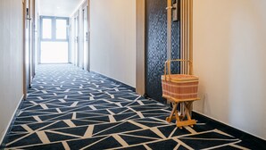 Un couloir d’hôtel avec un tapis à motifs, une chaise en bois et une porte donnant sur un mur à texture.