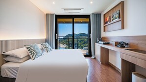 Une chambre d’hôtel moderne dotée d’un grand lit, d’un bureau en bois et d’un balcon offrant une vue sur les collines.