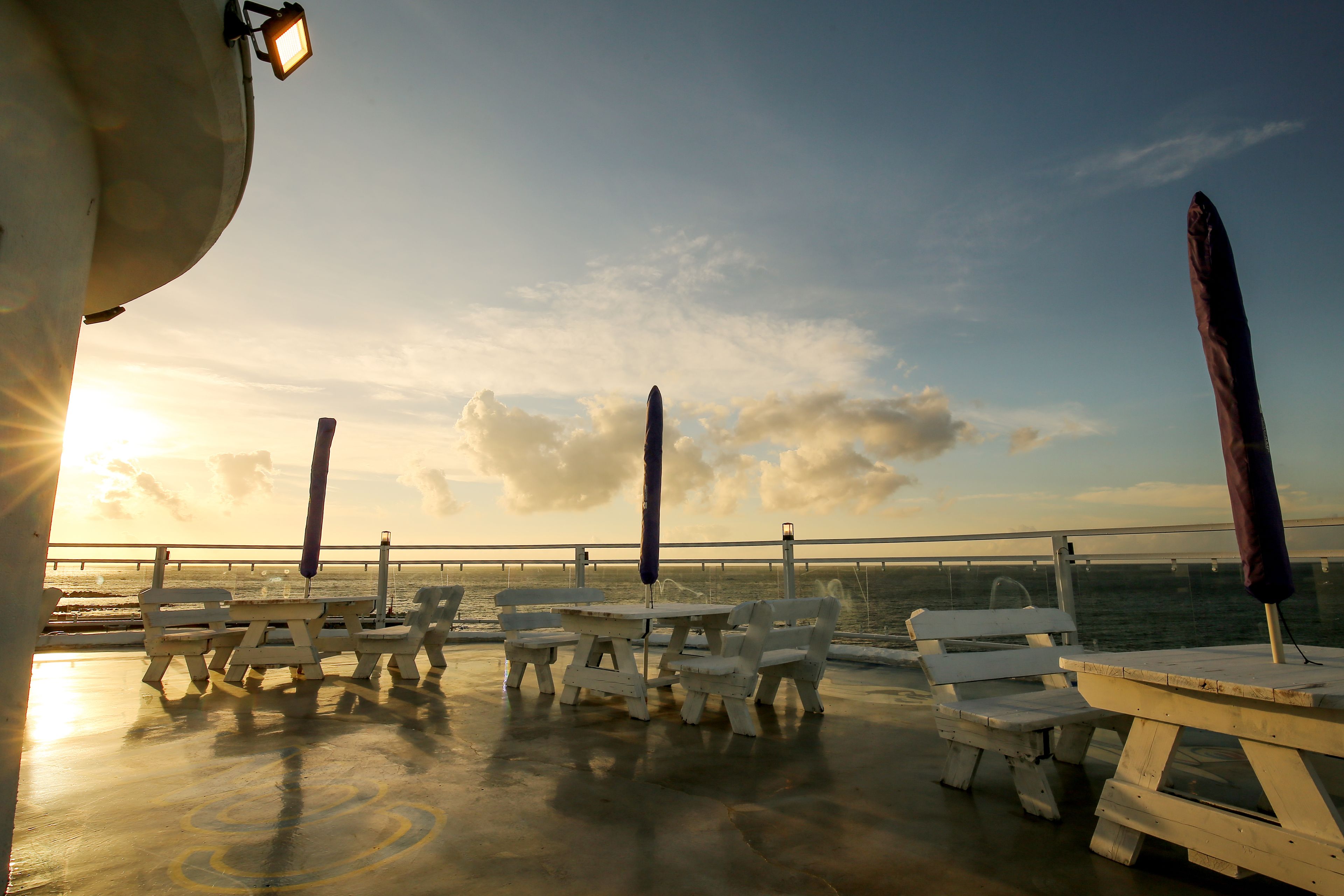Une terrasse avec des tables de pique-nique et des bancs donnant sur une étendue d’eau au coucher du soleil.