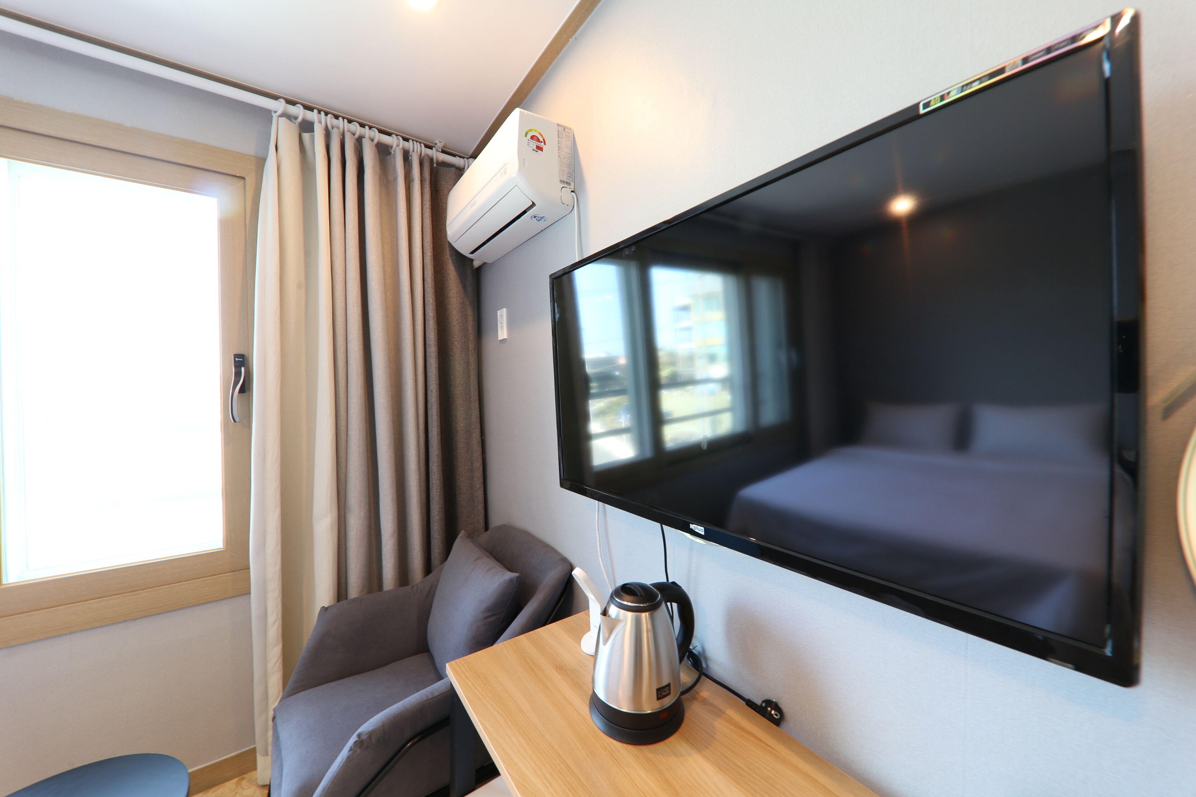 Ein modernes Hotelzimmer mit großem Flachbildfernseher an der Wand, einer Couch, einem kleinen Tisch mit Wasserkocher und einer Klimaanlage.