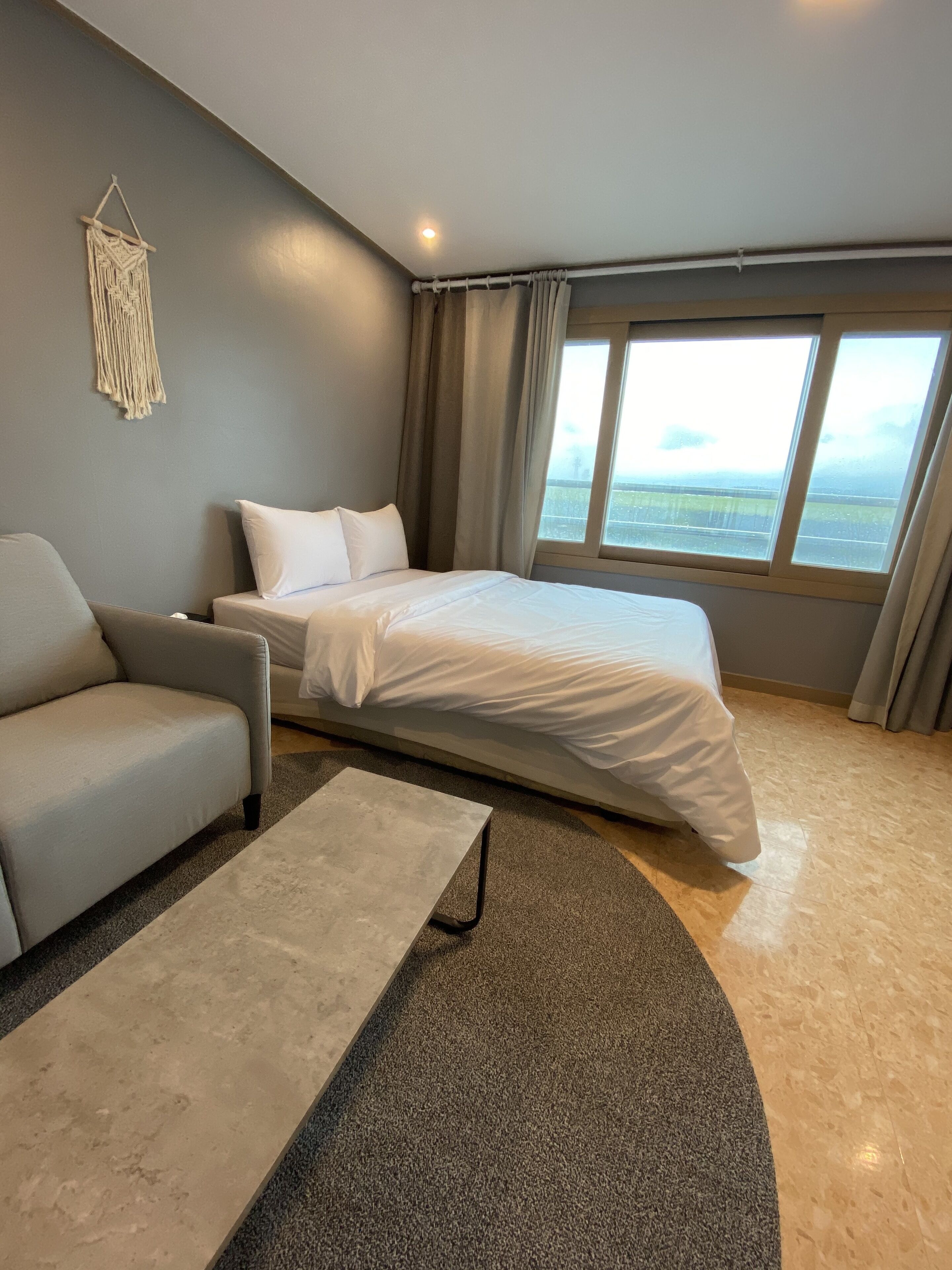 A modern hotel room with a bed, a sofa, a coffee table, and a view of the landscape through large windows.