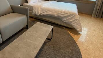 A modern hotel room with a bed, a sofa, a coffee table, and a view of the landscape through large windows.
