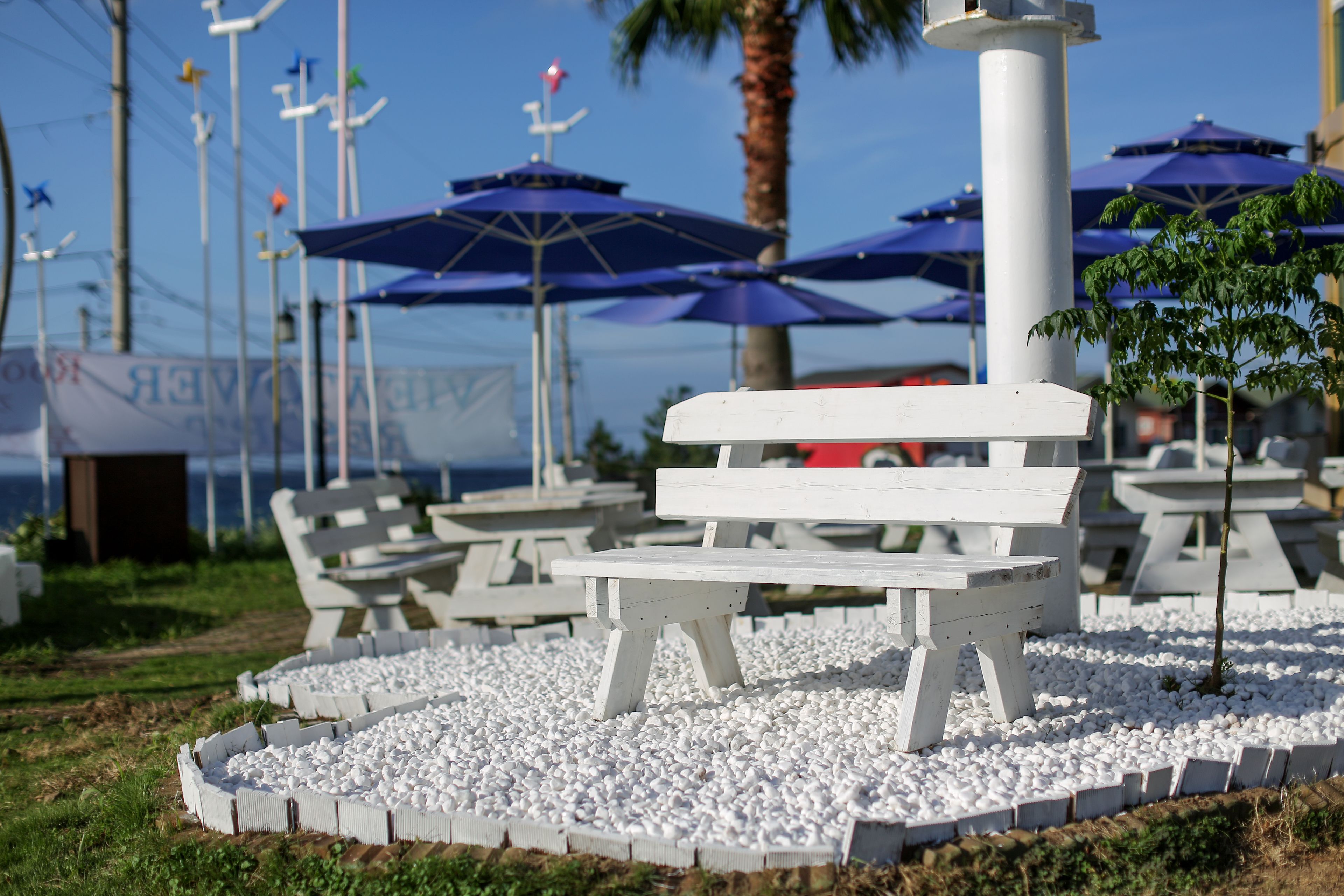 Outdoor seating with white benches and blue umbrellas, surrounded by white pebbles.