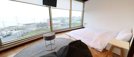 A bedroom with a large window, a bed, a small stool, and a television mounted on the wall.