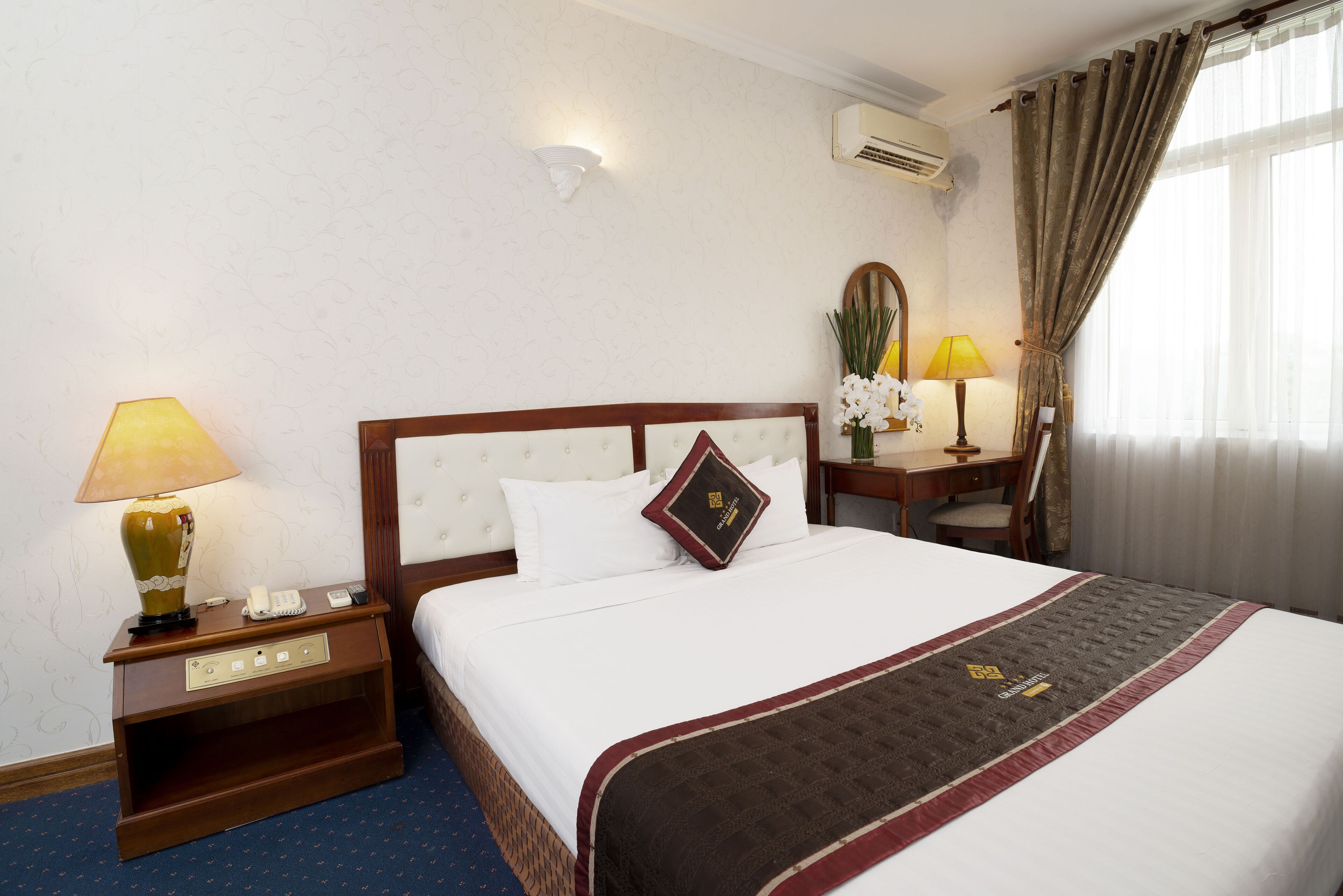 A hotel room with a large bed, a nightstand with a lamp, a desk with a chair, a mirror, and a window with curtains.