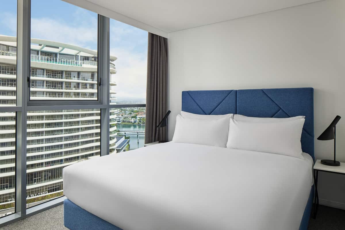 A modern hotel room with a large bed, a nightstand, and a view of a high-rise building and water.