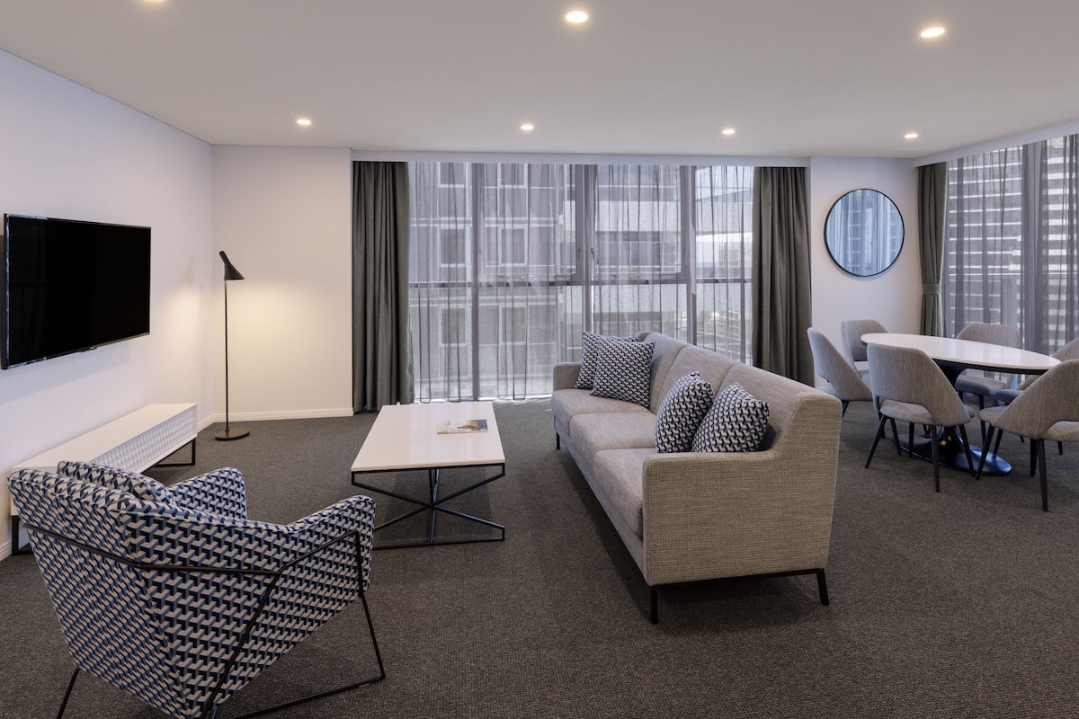 A modern hotel room with a sofa, armchair, dining table, and TV.