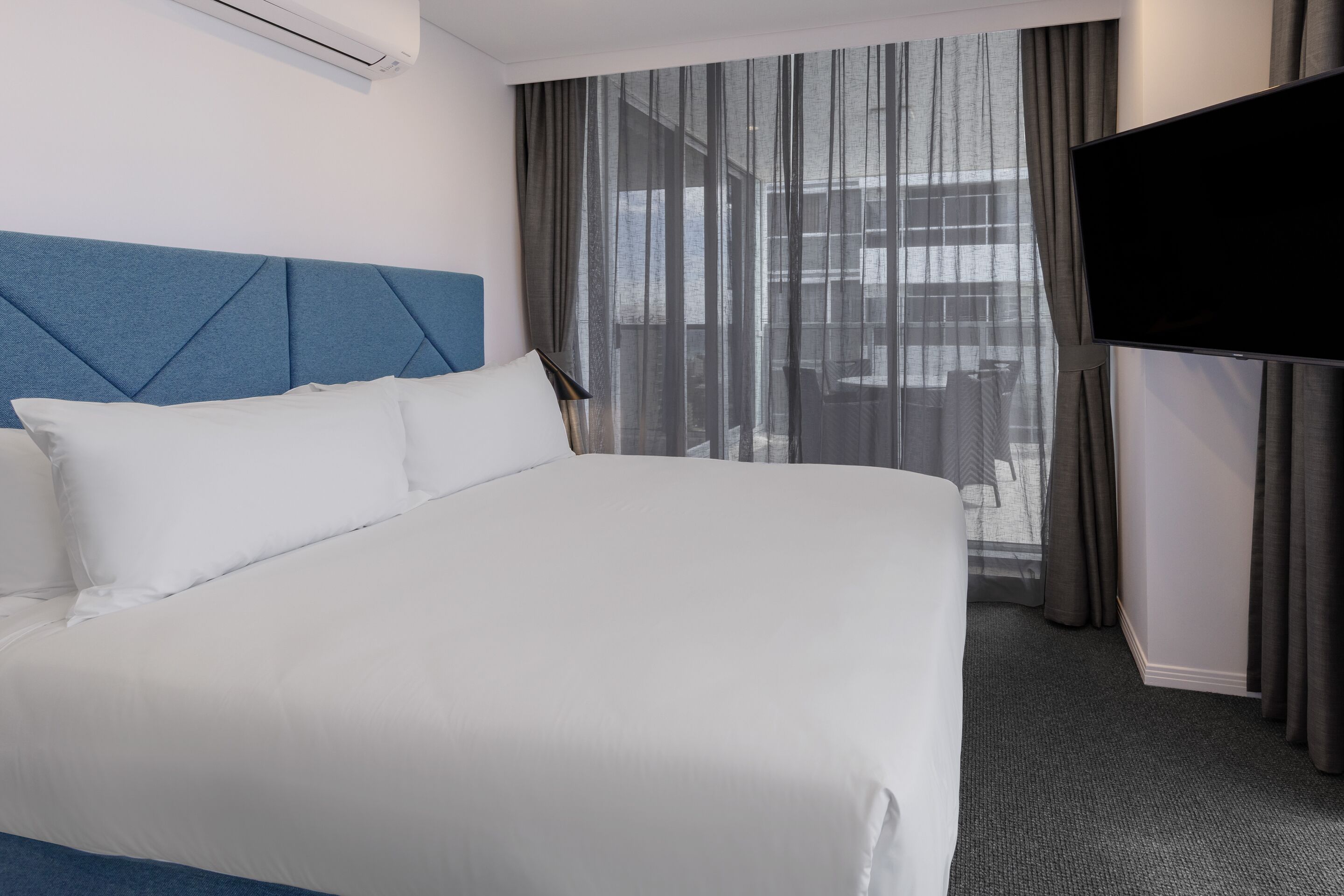 Une chambre d’hôtel avec un grand lit, un téléviseur à écran plat et une vue sur la ville à travers de fines rideaux.