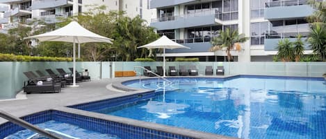 Una piscina con sillones de descanso y sombrillas, rodeada por un edificio de varios pisos con balcones.