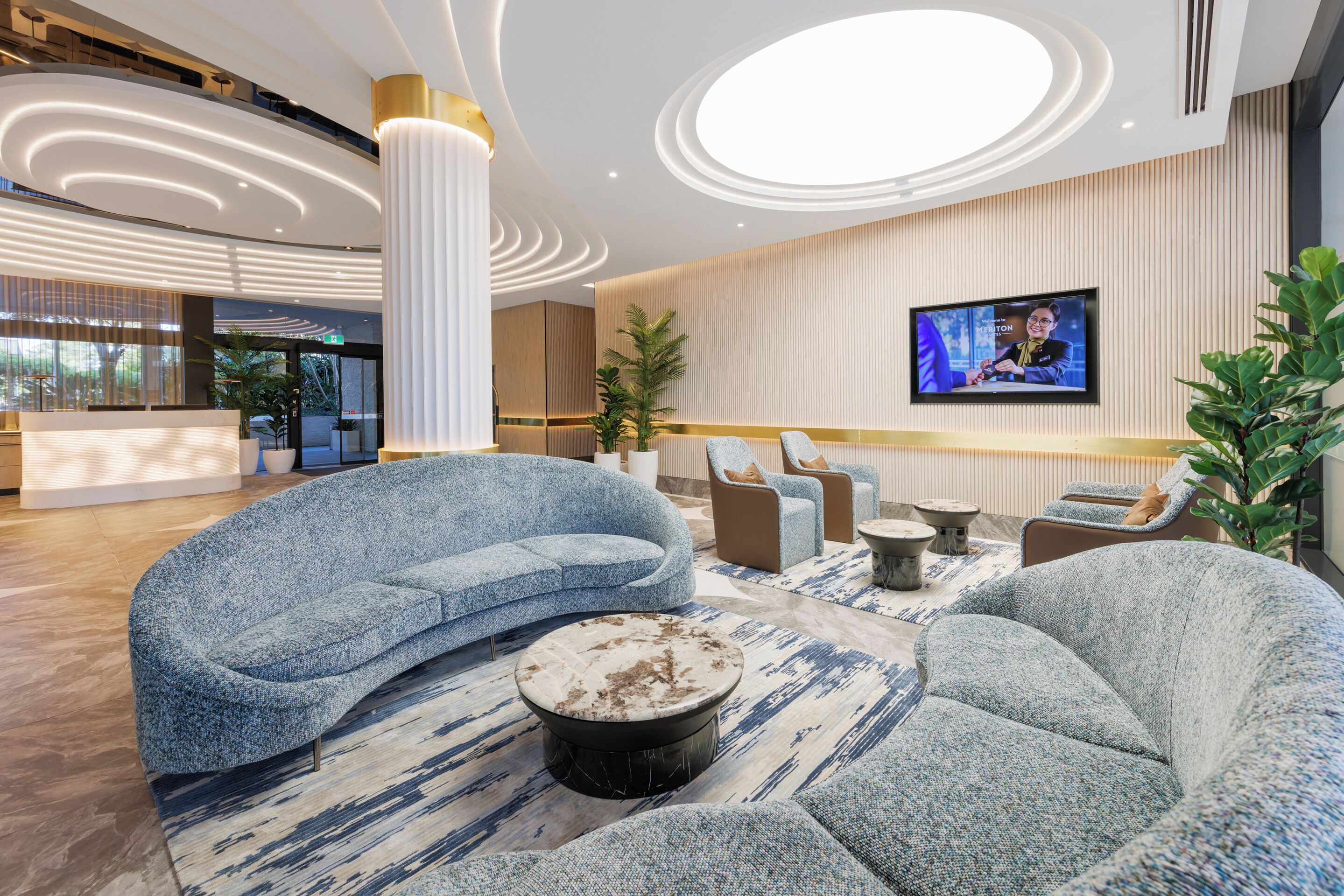 A modern lobby with a curved blue sofa, a round coffee table, and a TV screen displaying a person.