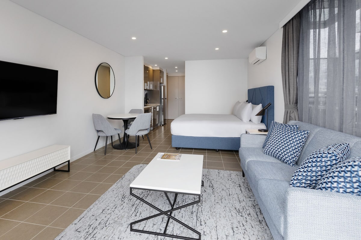A modern hotel room with a large bed, a sofa, a TV, a dining area, and a kitchen.