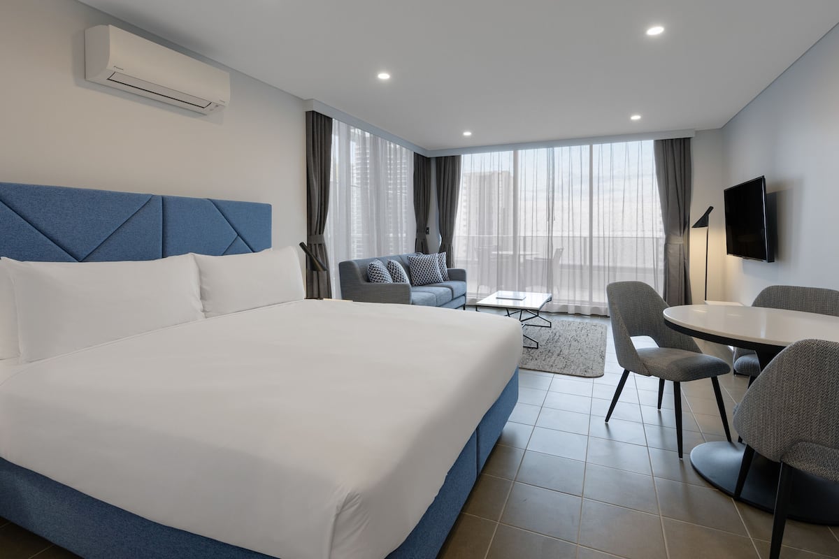 A hotel room with a large bed, a sofa, a dining table with chairs, and a TV mounted on the wall.