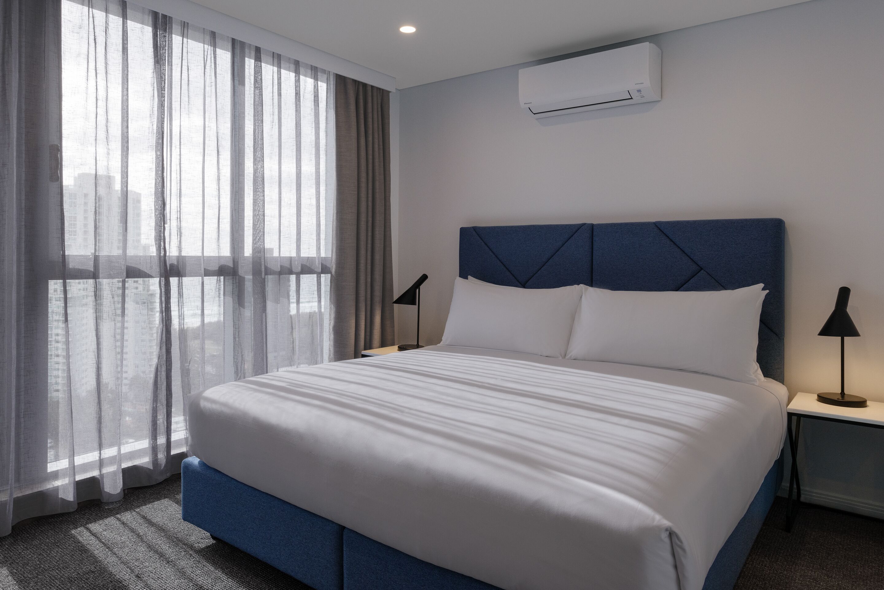 A hotel room with a large bed, a window with sheer curtains, a wall-mounted air conditioner, and a nightstand with a lamp.