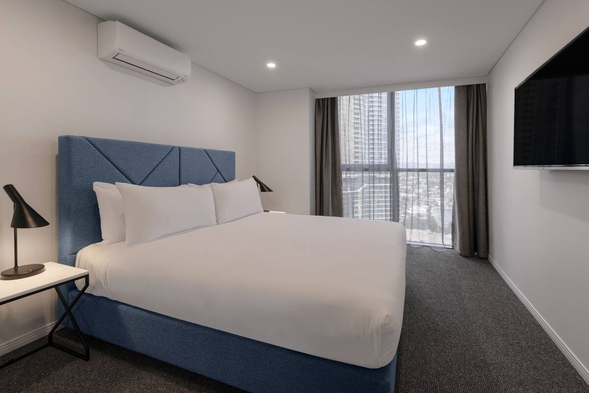 A modern hotel room with a large bed, a bedside table with a lamp, an air conditioning unit, a large window with curtains, and a flat-screen TV mounted on the wall.