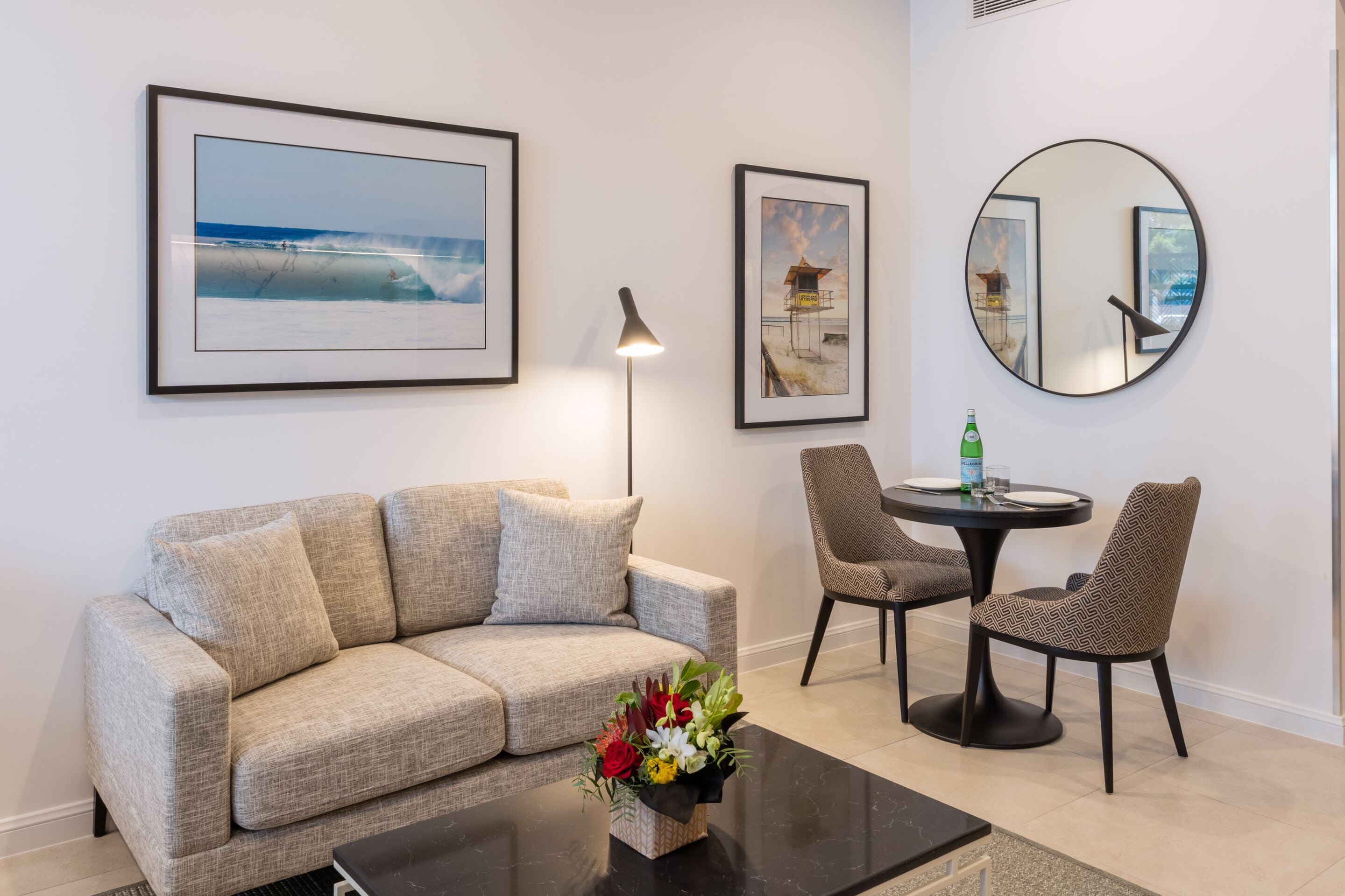 Sala de estar com sofá, mesa de centro com arranjo floral, mesa redonda com duas cadeiras, quadros na parede e espelho redondo.