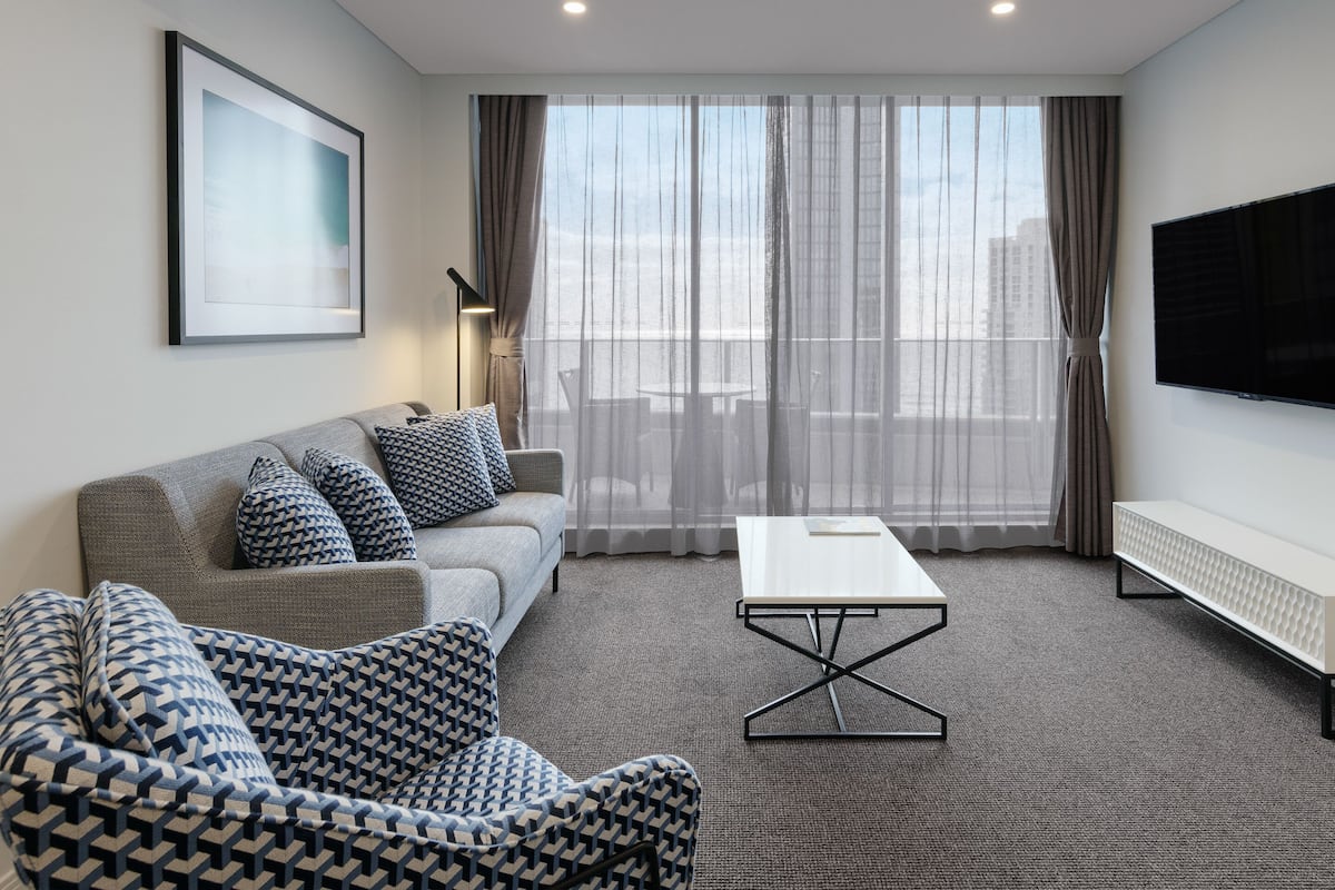 A modern living room with a grey sofa, patterned armchair, and a white coffee table. A flat-screen TV is mounted on the wall, and a large window with sheer curtains is visible.
