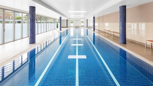 A long indoor swimming pool with blue tiles and white lane markings, surrounded by walls with blue and beige tiles, and a bench on the right side.