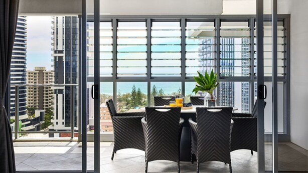 A modern dining area with a table and chairs, large windows with blinds, and a view of the cityscape and ocean.