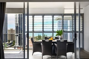 A modern dining area with a table and chairs, large windows with blinds, and a view of the cityscape and ocean.