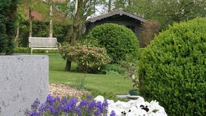 Un jardín con un banco, un cobertizo pequeño y diversas plantas, incluidas flores y un arbusto bien cuidado.