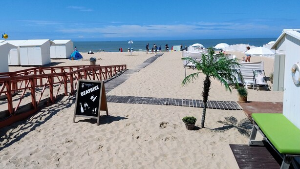 Zona de playa con cabañas blancas, una pasarela de madera y un cartel.
