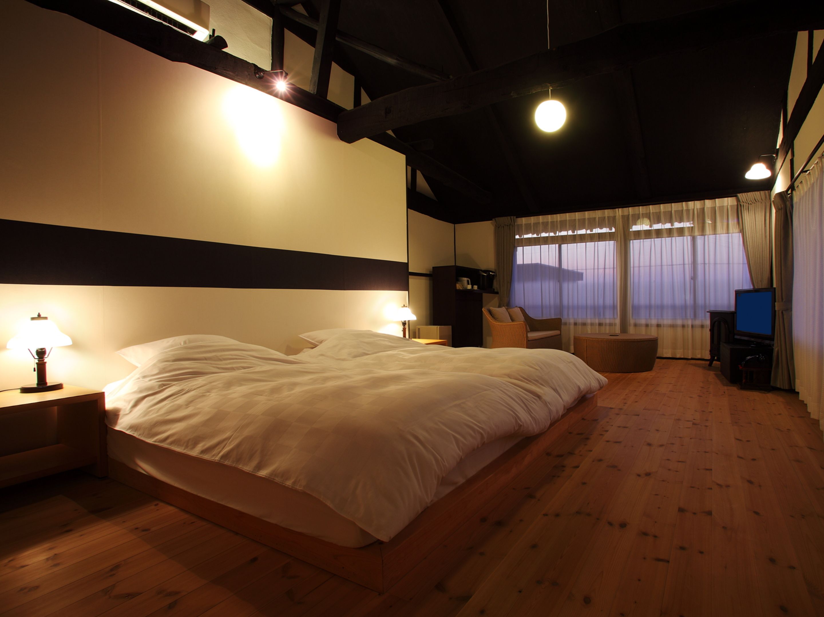 Ein Schlafzimmer mit einem großen Bett, zwei Nachttischen mit Lampen, einem Sessel, einem Fernseher und einem Fenster mit Vorhängen.
