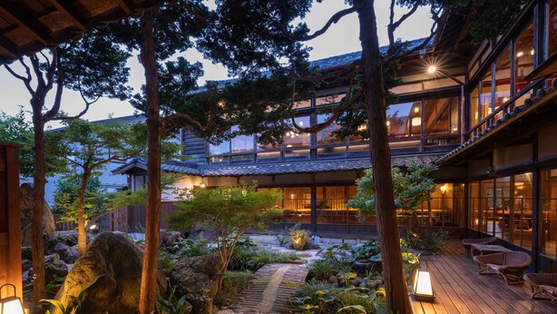 Un bâtiment de style japonais traditionnel doté d’une terrasse en bois, d’un chemin en pierre et d’un espace extérieur aménagé pour s’asseoir.