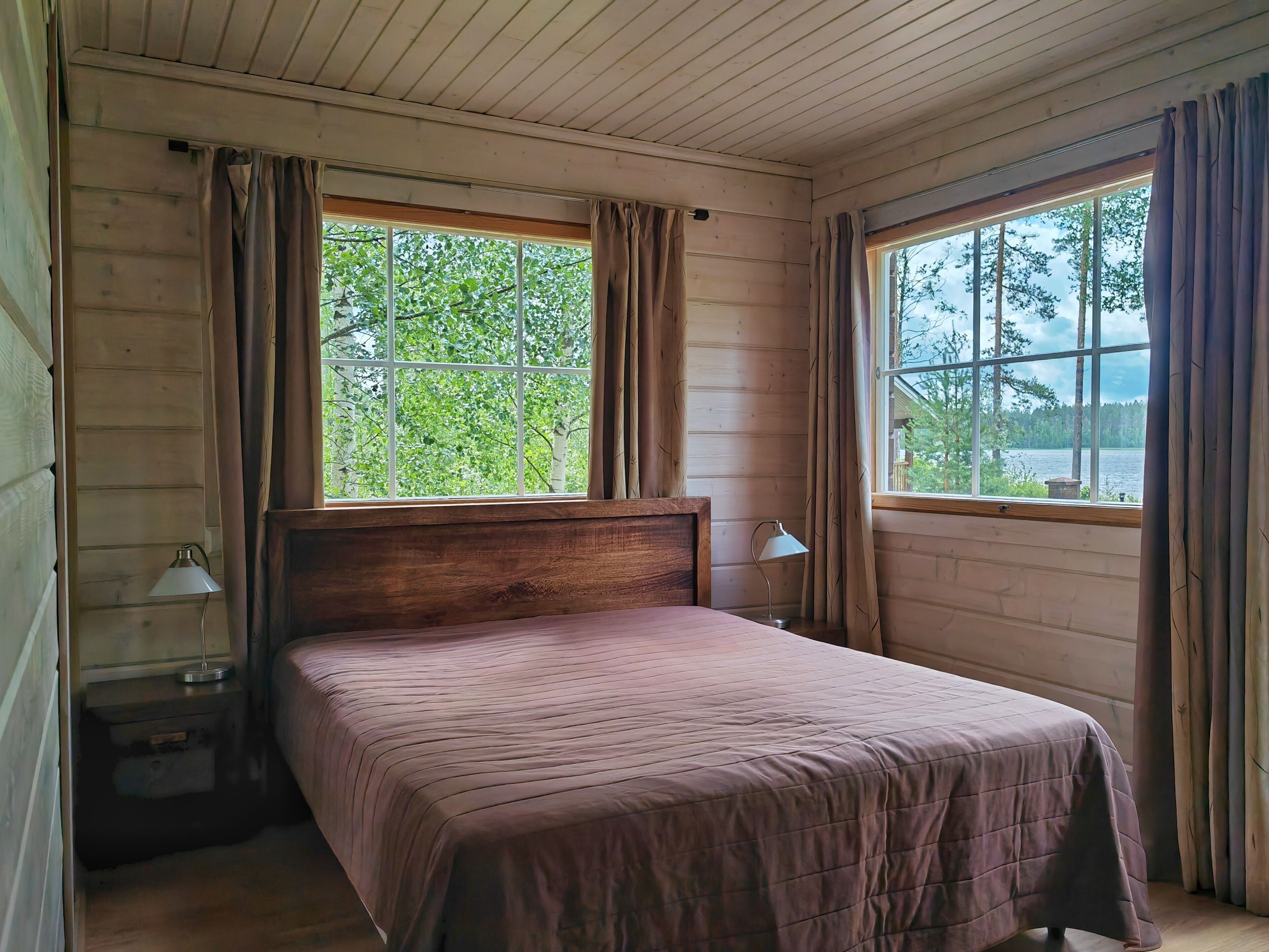 Quarto com cama de madeira, duas luminárias de cabeceira e janelas amplas com cortinas.