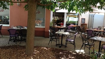 Un café en plein air avec des tables et des chaises, un arbre et un bâtiment en arrière-plan.