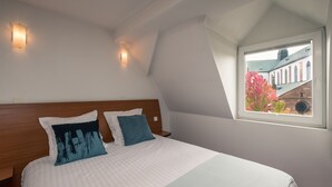 A bedroom with a large bed, two wall sconces, a window with a view of buildings and trees, and a sloped ceiling. - Notre Dame (Haguenau)
