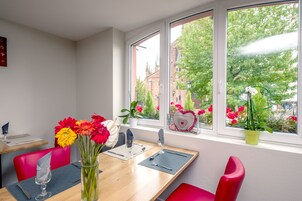 Un comedor con una mesa preparada para dos personas, un florero con flores y una ventana con vistas a edificios y árboles.