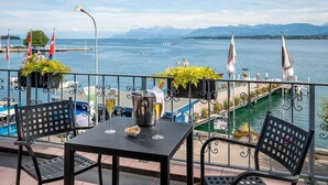 Une terrasse avec une table, des chaises et des plantes en pot, donnant sur un lac où l’on peut voir des bateaux et un pont.