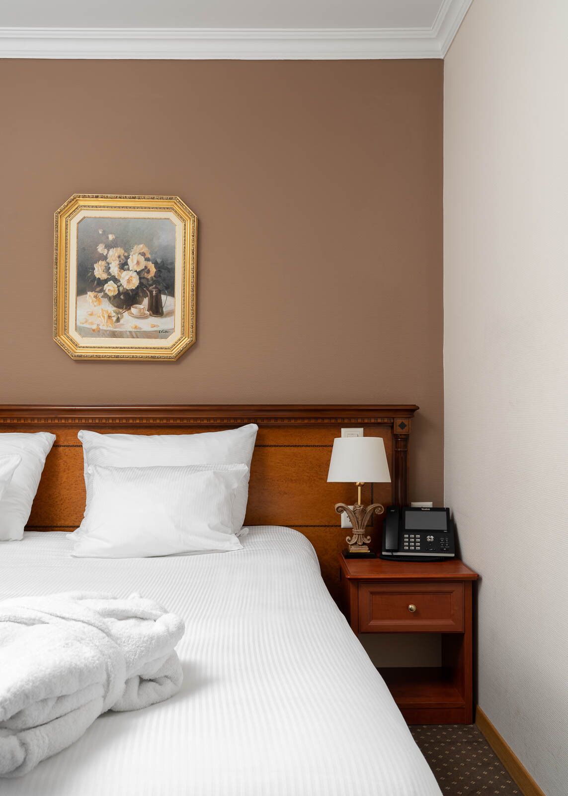 A hotel room with a large bed, a nightstand with a lamp and phone, and a framed painting on the wall.