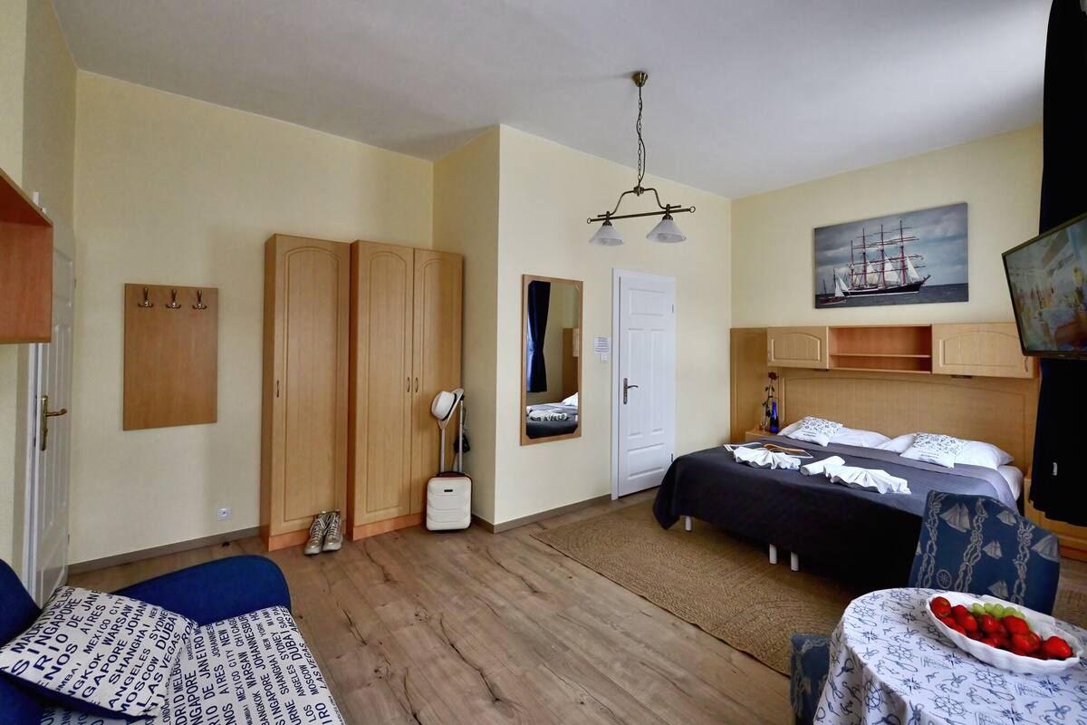A room with two beds, a desk with a chair, a small table with fruit, a mirror, and a ship painting on the wall.