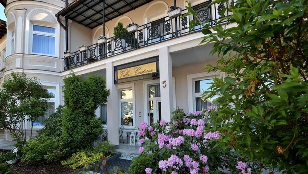 Un hotel con balcón, un letrero que dice "Villa Castoria" y un jardín con flores rosadas.