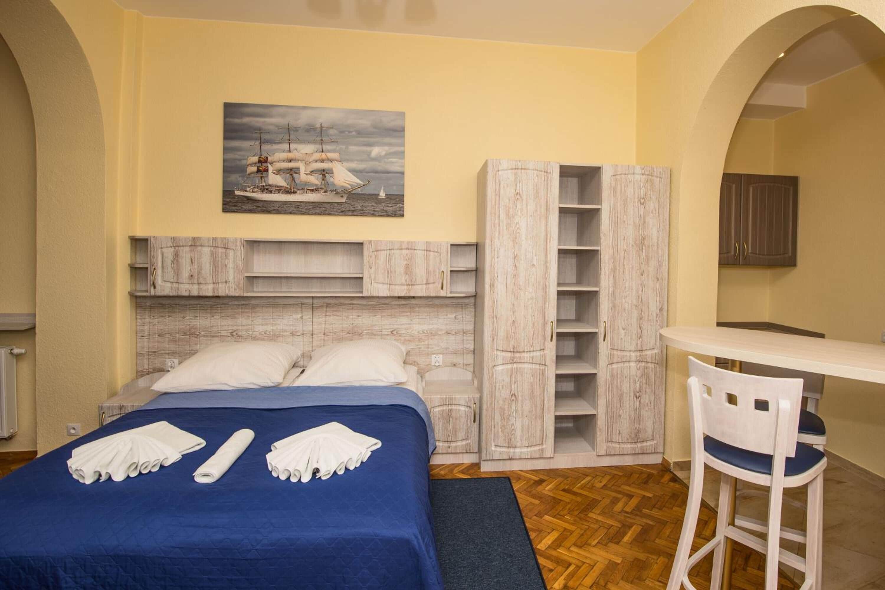 A bedroom with a bed, a desk, and a chair. There is a painting on the wall and a wardrobe.