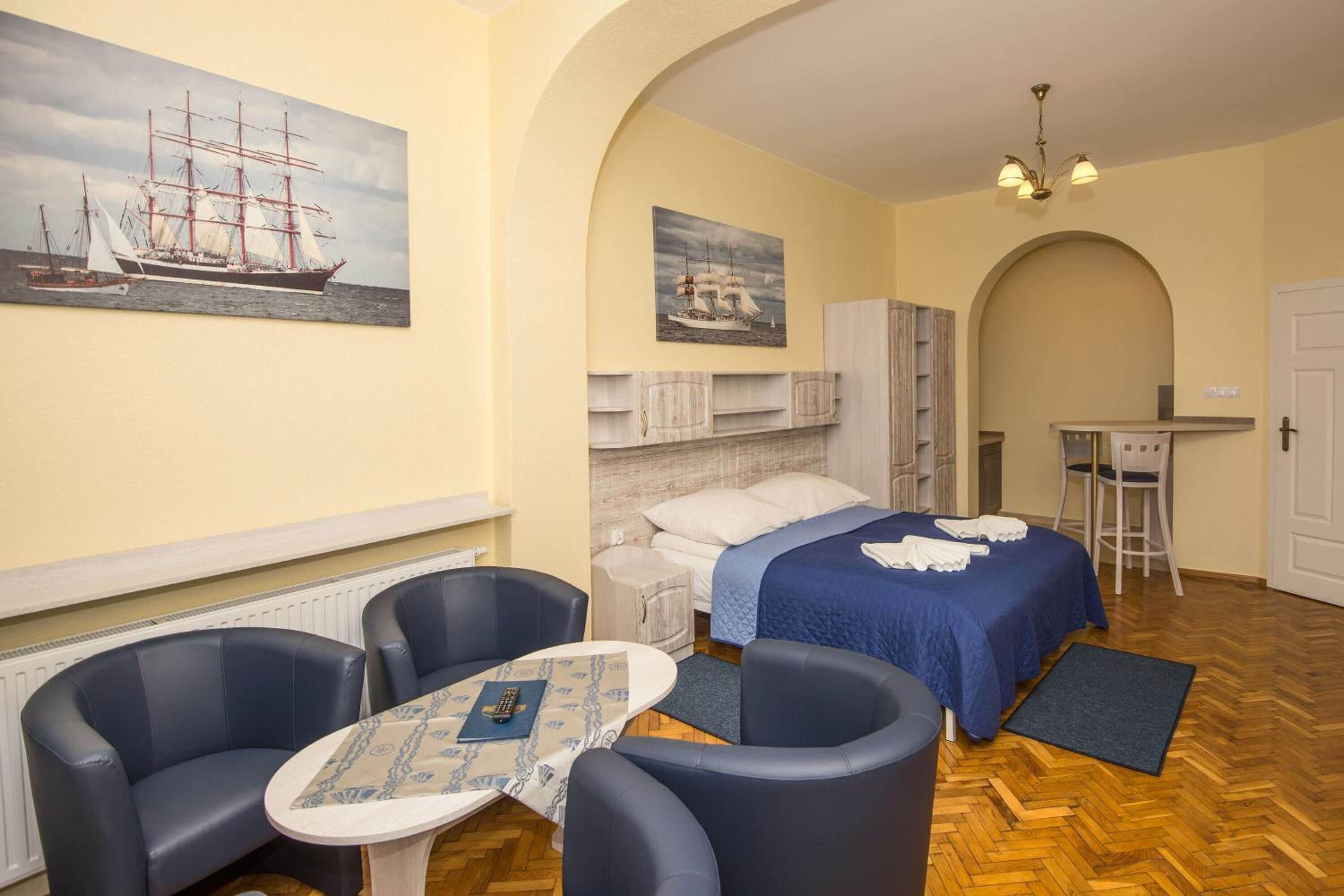 A hotel room with a bed, two chairs, a small table, a desk, and a painting of a ship on the wall.