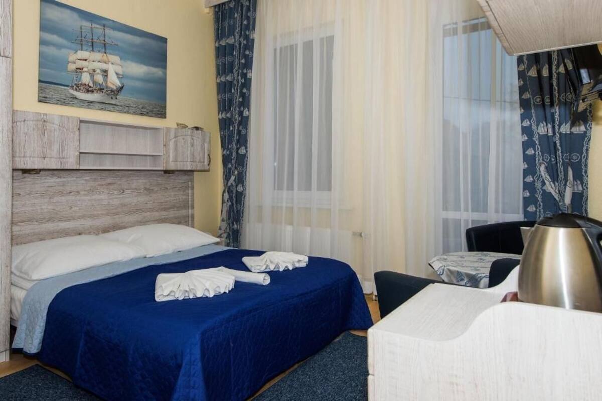 A hotel room with a bed, a desk, a chair, a window with sheer curtains, and a painting of a ship on the wall.