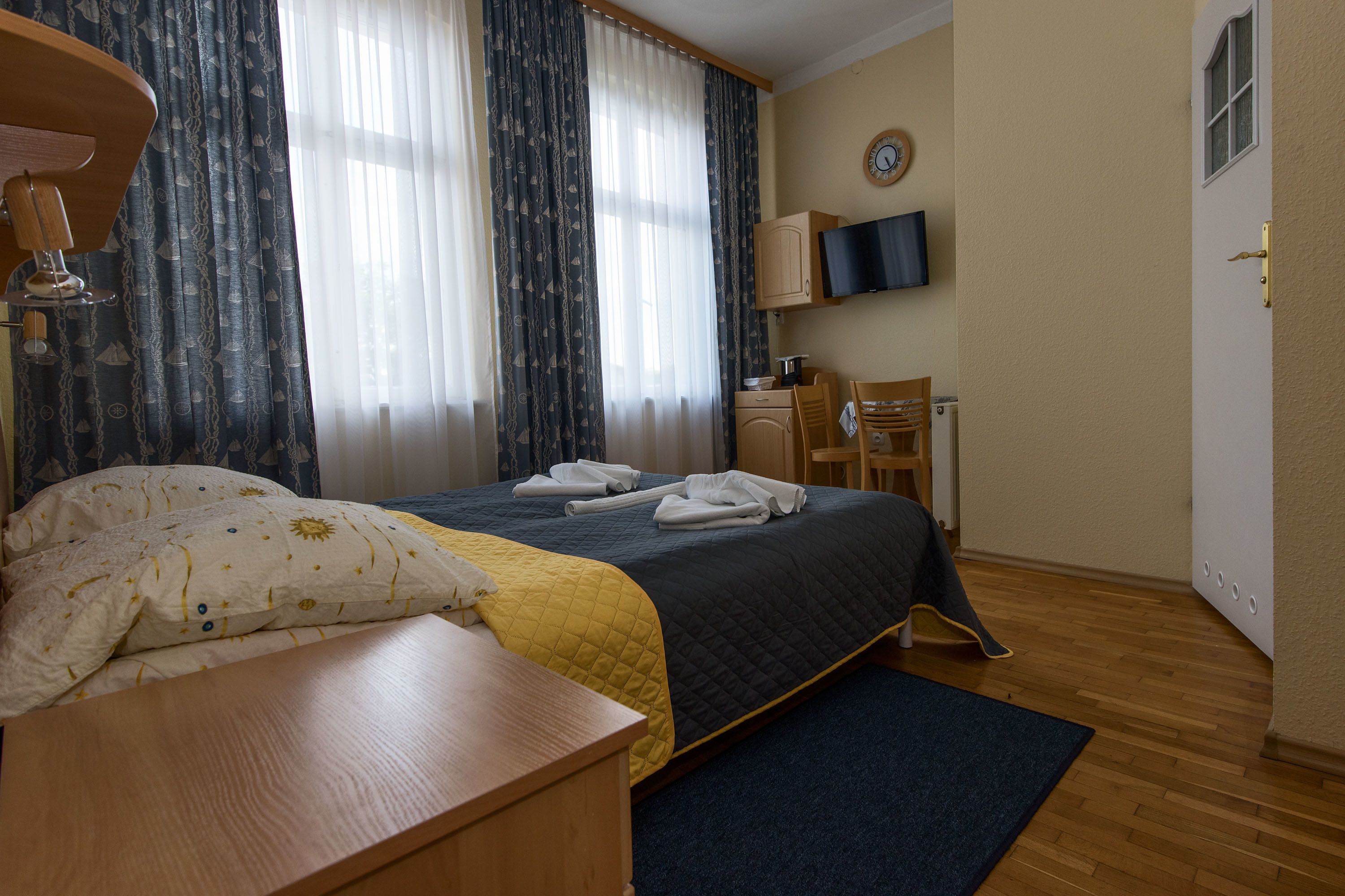 A hotel room with a bed, a desk, a chair, a TV, and a clock on the wall.