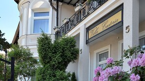 Un edificio con balcón, un letrero que dice 'Villa Caria' y un jardín con flores rosadas.