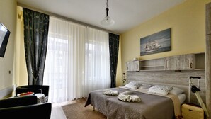 Habitación de hotel ordenada con cama, escritorio, silla, mesa de comedor con frutero y una ventana grande con cortinas.