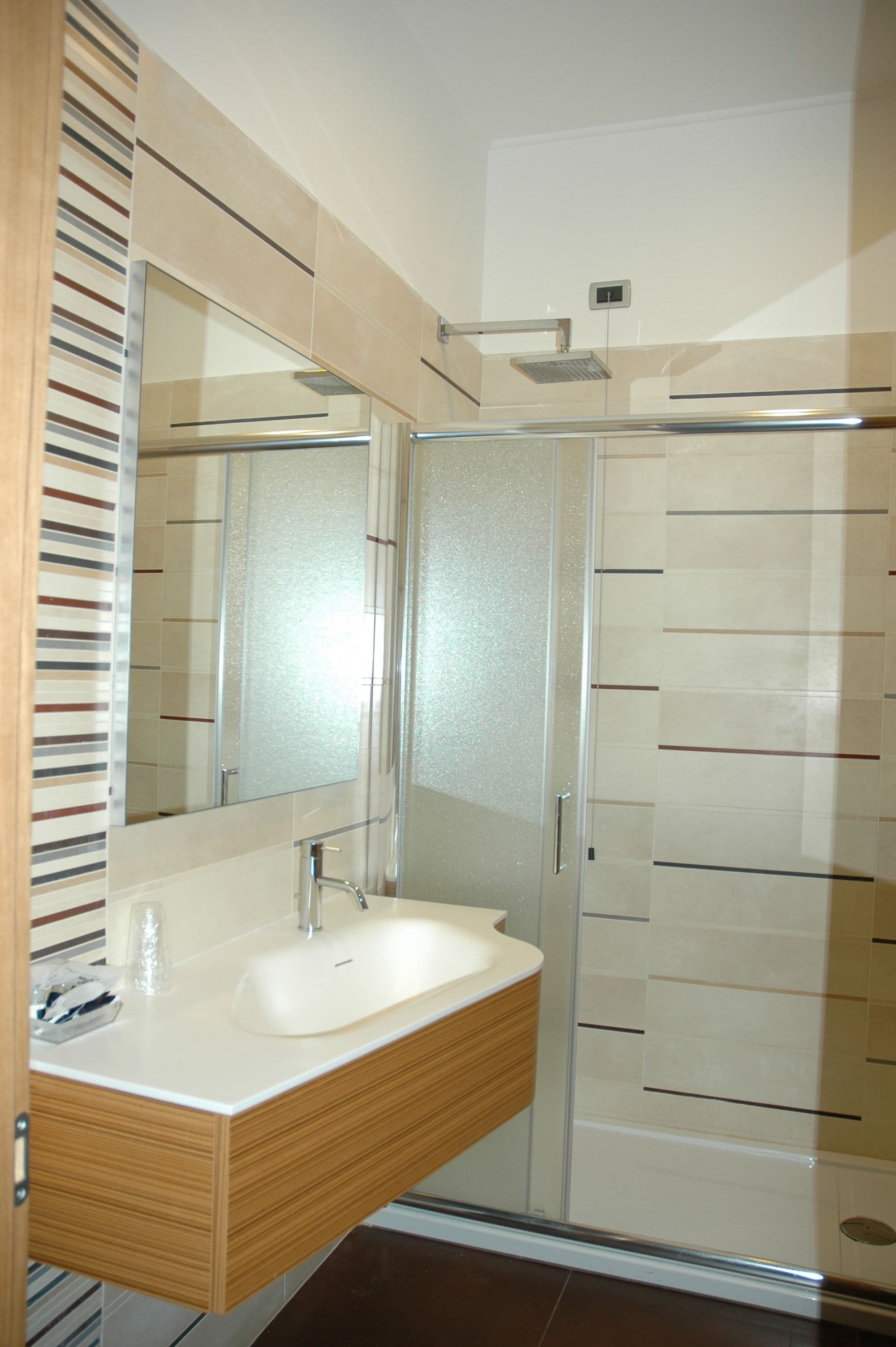 A modern bathroom with a wooden vanity, a large mirror, a glass shower enclosure, and a tiled shower wall.