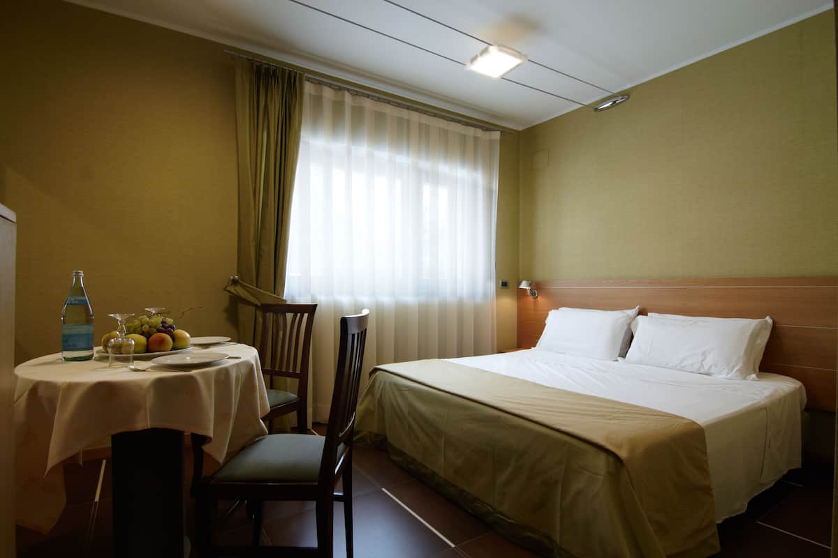 Habitación de hotel con cama, una mesa con botella y fruta, una silla y una ventana con cortinas.