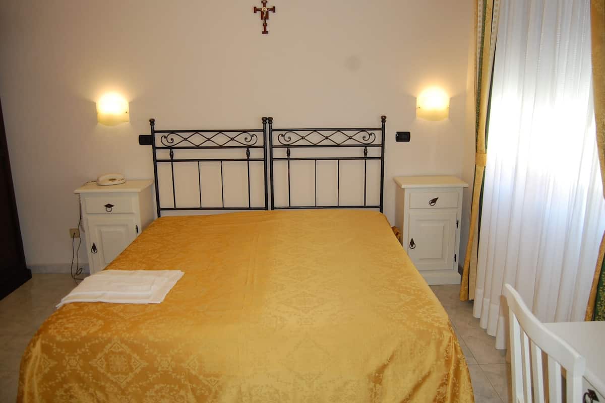 Un dormitorio con cama, dos mesitas de noche, una mesilla auxiliar, una cruz en la pared y una ventana con cortinas.