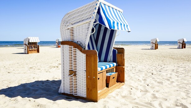 Ein Liegestuhl mit einem gestreiften Baldachin, Holzrahmen und gepolstertem Sitz, auf einem Sandstrand mit dem Meer im Hintergrund.