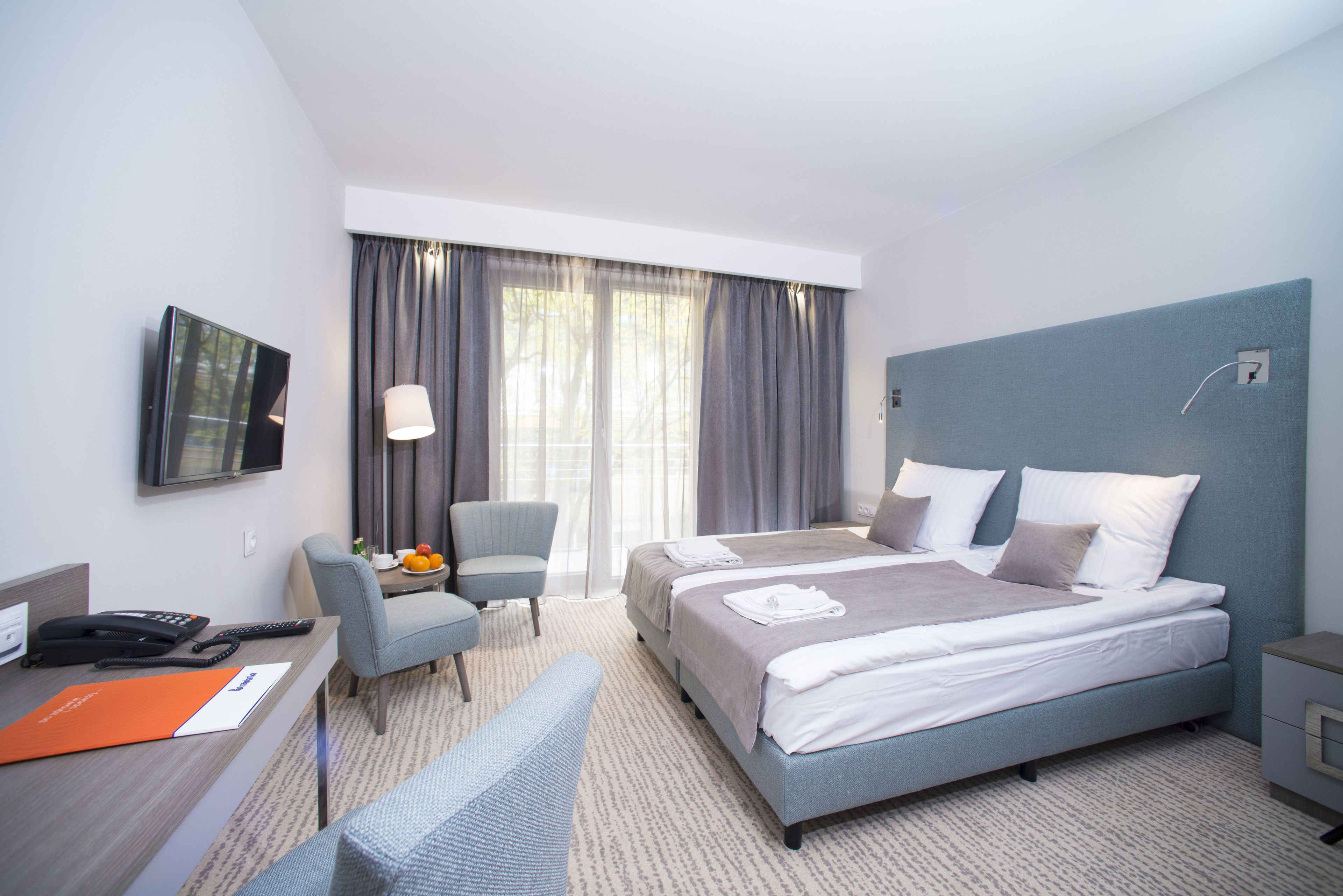 Une chambre d’hôtel moderne dotée d’un grand lit, d’un bureau avec un téléphone, d’une chaise, d’un téléviseur fixé au mur et d’une fenêtre avec des rideaux.