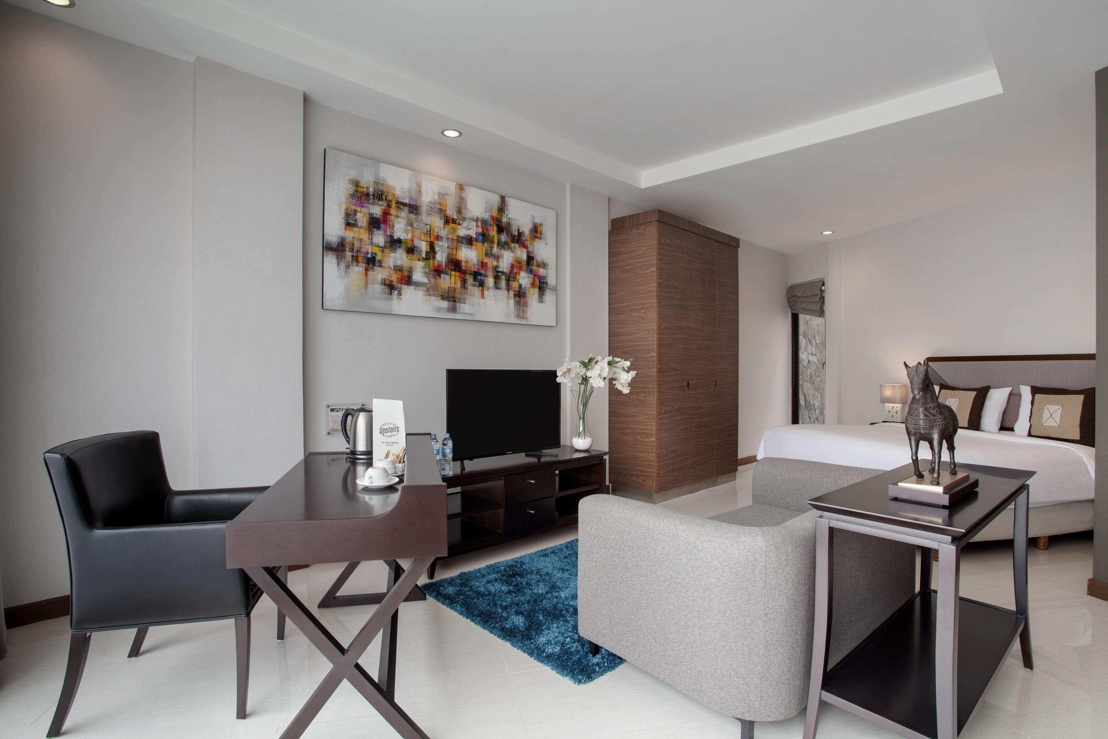 A modern hotel room with a flat-screen TV, a desk with a chair, a sofa, a bed, a nightstand, and a large abstract painting on the wall.