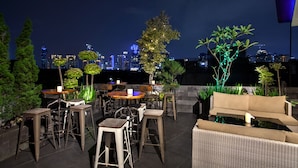 An outdoor seating area with tables and chairs, surrounded by potted plants and trees, overlooking a city skyline at night. - Sawana Suites (Jakarta)