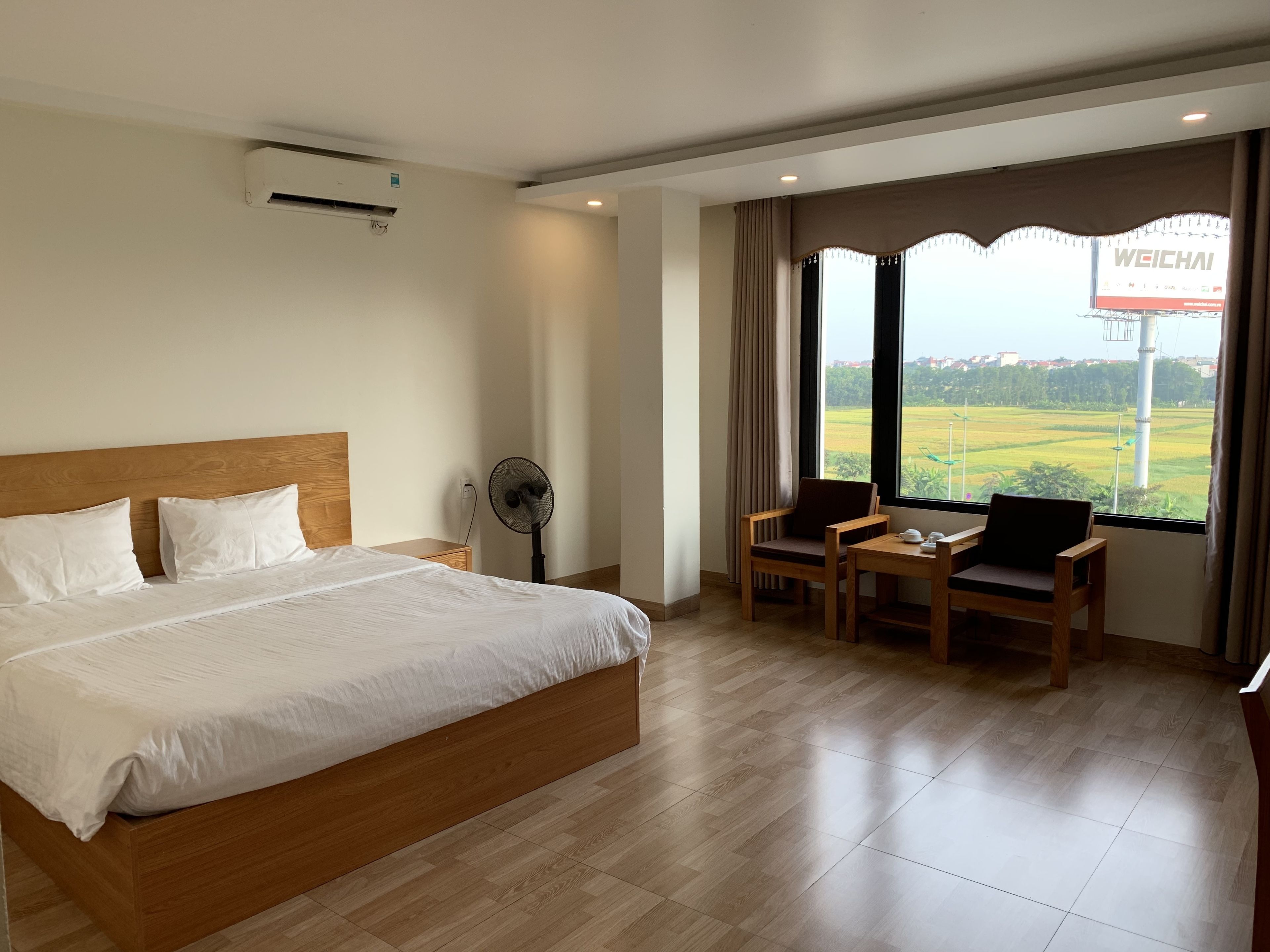 A hotel room with a large bed, a desk with two chairs, a standing fan, and a view of a field through the window.